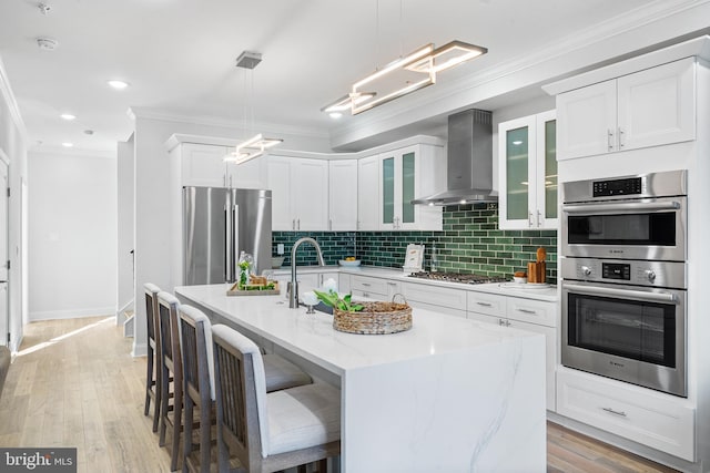 kitchen featuring light stone countertops, wall chimney range hood, decorative light fixtures, a kitchen island with sink, and appliances with stainless steel finishes