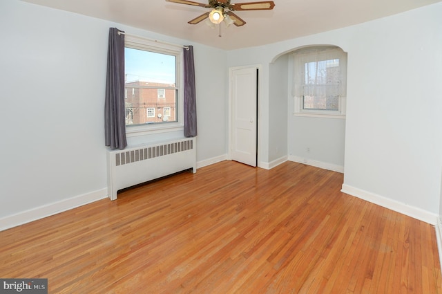 unfurnished room featuring ceiling fan, arched walkways, light wood-style flooring, baseboards, and radiator