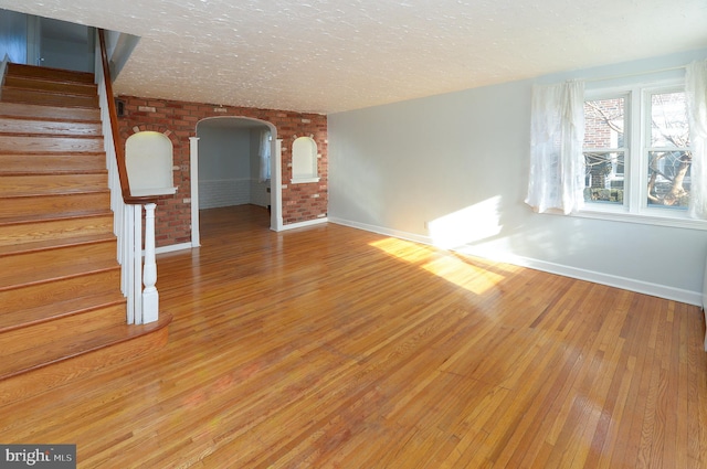 unfurnished living room with light wood finished floors, arched walkways, brick wall, stairs, and a textured ceiling