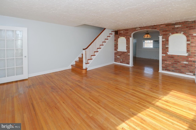 unfurnished living room with arched walkways, stairway, a textured ceiling, brick wall, and wood finished floors