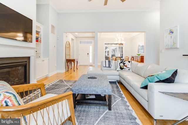 living room featuring built in shelves, light wood-type flooring, ceiling fan with notable chandelier, and ornamental molding