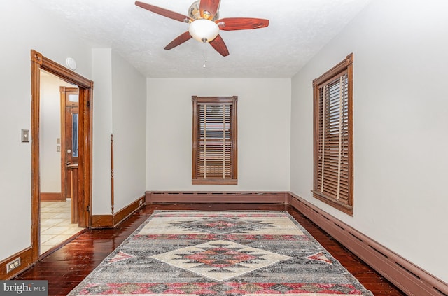 empty room with a textured ceiling, ceiling fan, dark wood-type flooring, and a baseboard heating unit