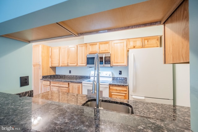 kitchen with light brown cabinetry, dark stone counters, and white appliances