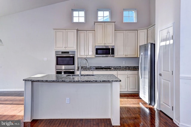 kitchen with sink, stainless steel appliances, dark hardwood / wood-style floors, dark stone countertops, and an island with sink