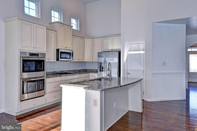 kitchen with a towering ceiling, dark stone counters, stainless steel appliances, sink, and a center island with sink