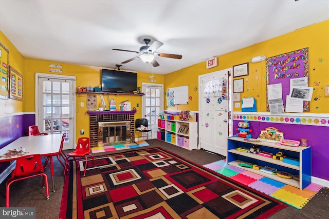 recreation room with ceiling fan, dark colored carpet, and a brick fireplace