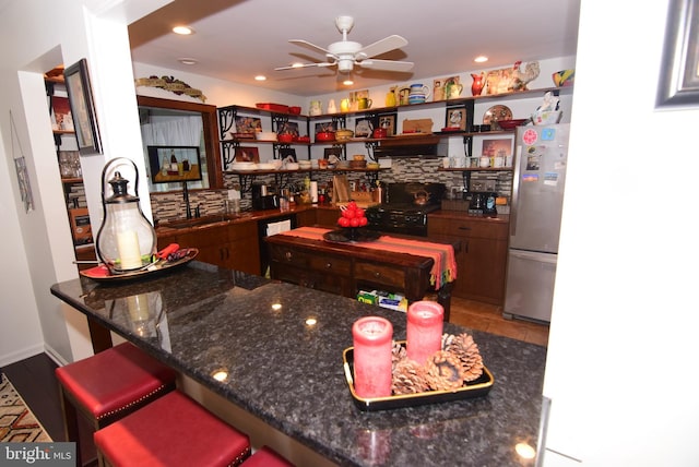 kitchen featuring ceiling fan, sink, dark stone countertops, stainless steel fridge, and a breakfast bar area