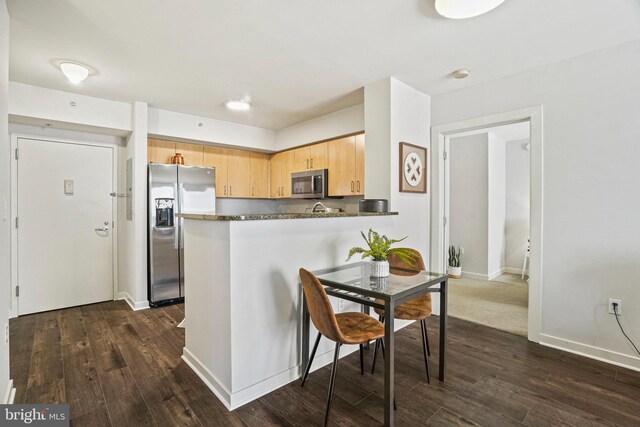 kitchen with light brown cabinets, dark hardwood / wood-style flooring, kitchen peninsula, dark stone countertops, and appliances with stainless steel finishes