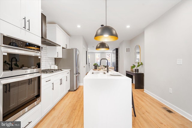 kitchen with white cabinetry, wall chimney exhaust hood, hanging light fixtures, an island with sink, and appliances with stainless steel finishes