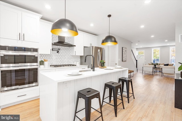 kitchen with appliances with stainless steel finishes, an island with sink, white cabinetry, and wall chimney range hood