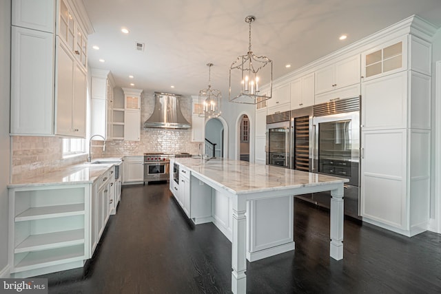 kitchen with a large island, wall chimney range hood, premium appliances, pendant lighting, and white cabinets