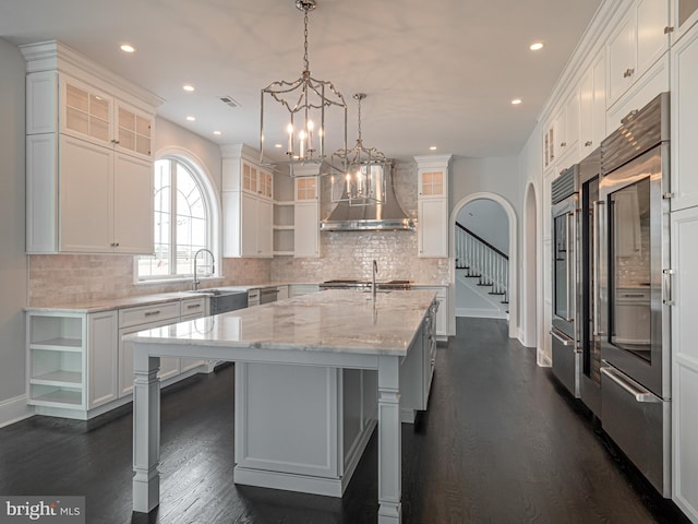 kitchen featuring light stone countertops, wall chimney exhaust hood, white cabinets, hanging light fixtures, and an island with sink