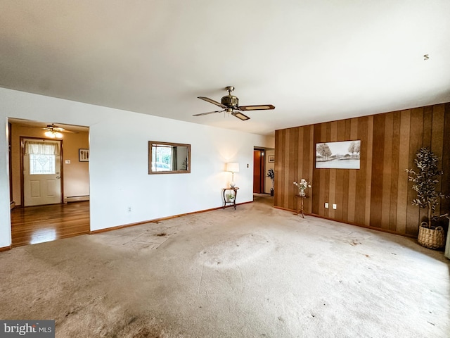 unfurnished living room with a baseboard radiator, wooden walls, a ceiling fan, baseboards, and carpet