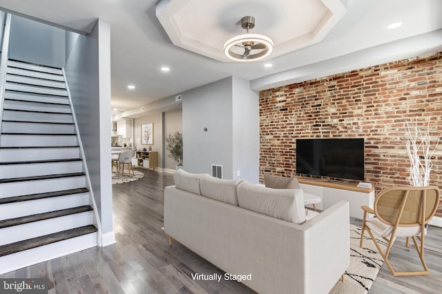 living room with a tray ceiling, stairway, wood finished floors, and brick wall