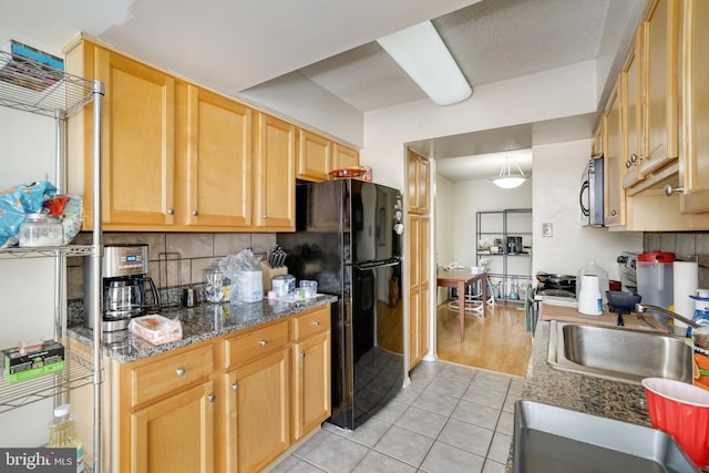 kitchen featuring light brown cabinets, dark stone counters, black fridge, sink, and light tile patterned flooring