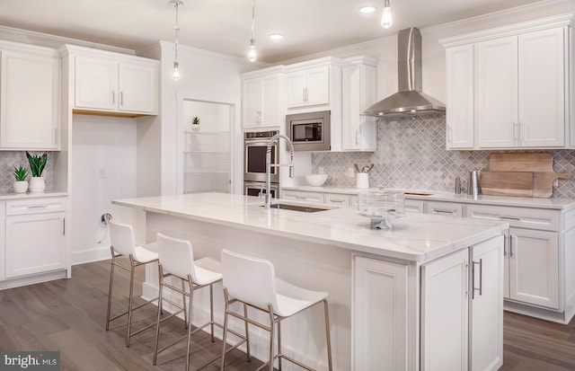 kitchen with white cabinetry, hanging light fixtures, stainless steel appliances, wall chimney range hood, and an island with sink