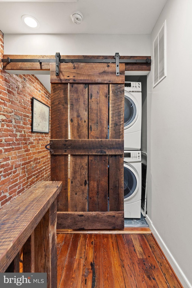 laundry room featuring a barn door, dark hardwood / wood-style flooring, brick wall, and stacked washer and clothes dryer