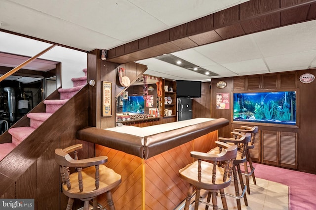 bar featuring refrigerator, light colored carpet, wooden walls, and a paneled ceiling