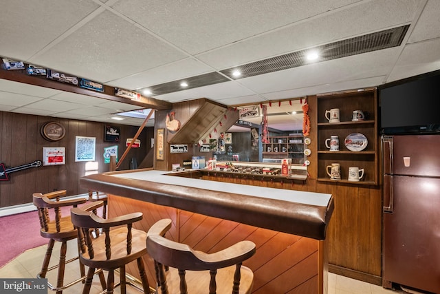 bar featuring a paneled ceiling, light tile patterned floors, stainless steel fridge, wooden walls, and built in shelves