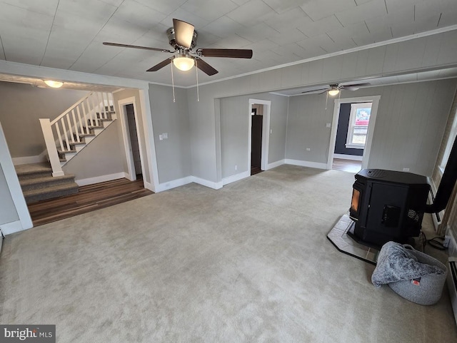 unfurnished living room featuring a wood stove, ceiling fan, carpet floors, and ornamental molding