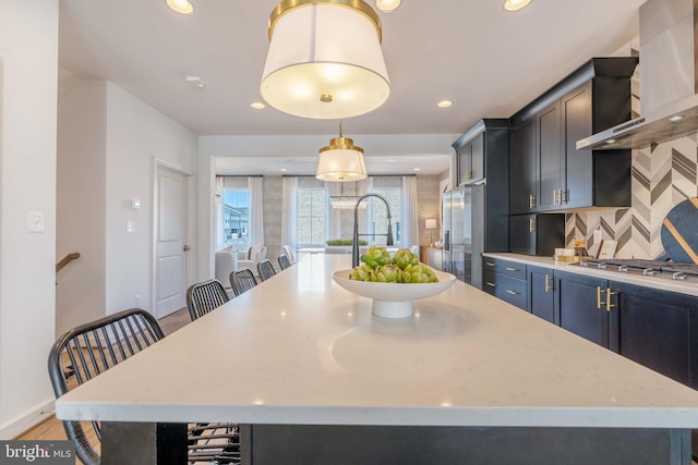 kitchen with a center island with sink, wall chimney range hood, hanging light fixtures, and tasteful backsplash