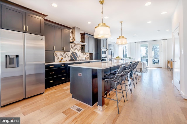 kitchen with wall chimney range hood, an island with sink, pendant lighting, a breakfast bar area, and appliances with stainless steel finishes