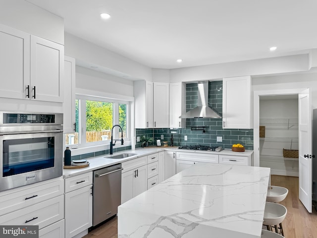 kitchen featuring a kitchen bar, appliances with stainless steel finishes, wall chimney range hood, sink, and white cabinetry