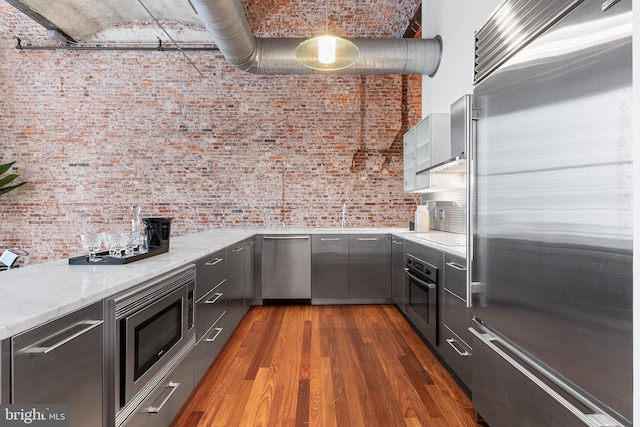 kitchen with built in appliances, brick wall, dark wood-style flooring, and light stone countertops