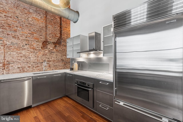 kitchen with brick wall, appliances with stainless steel finishes, dark wood-type flooring, wall chimney range hood, and a sink