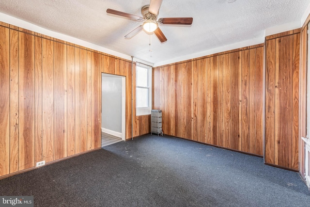 empty room with dark carpet, a textured ceiling, ceiling fan, wooden walls, and radiator heating unit