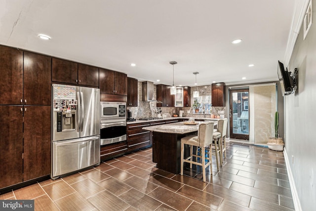 kitchen with a center island, wall chimney exhaust hood, hanging light fixtures, a breakfast bar, and appliances with stainless steel finishes