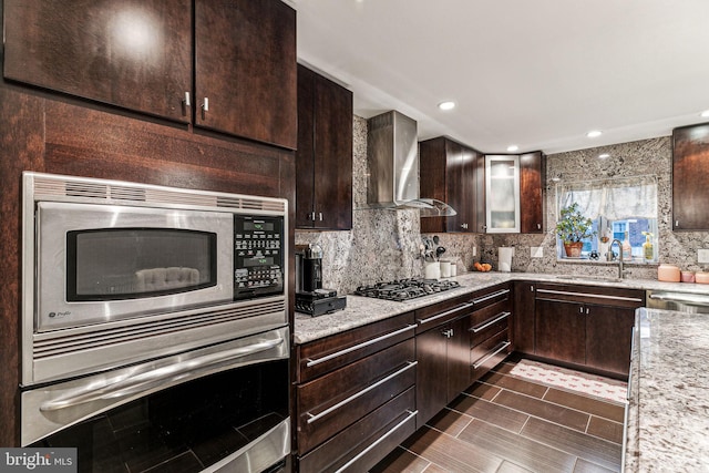 kitchen featuring light stone countertops, dark brown cabinets, stainless steel appliances, and wall chimney range hood