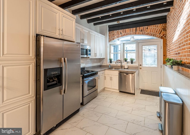 kitchen featuring ceiling fan, sink, beamed ceiling, brick wall, and appliances with stainless steel finishes