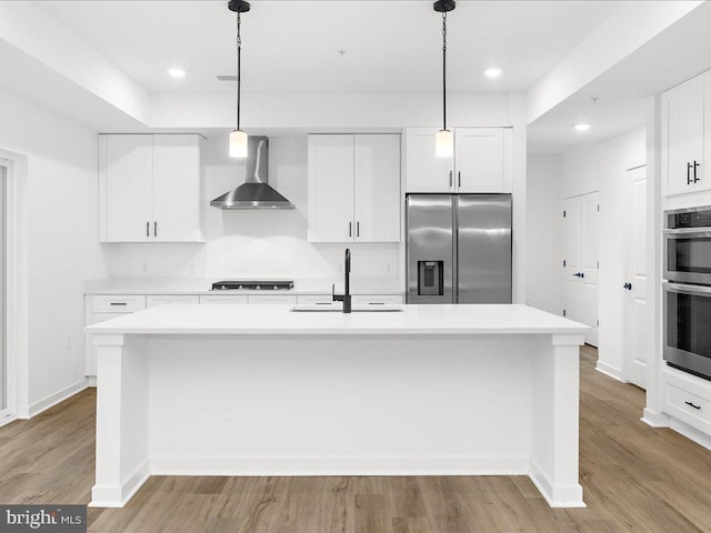 kitchen featuring stainless steel appliances, wall chimney range hood, pendant lighting, white cabinetry, and an island with sink