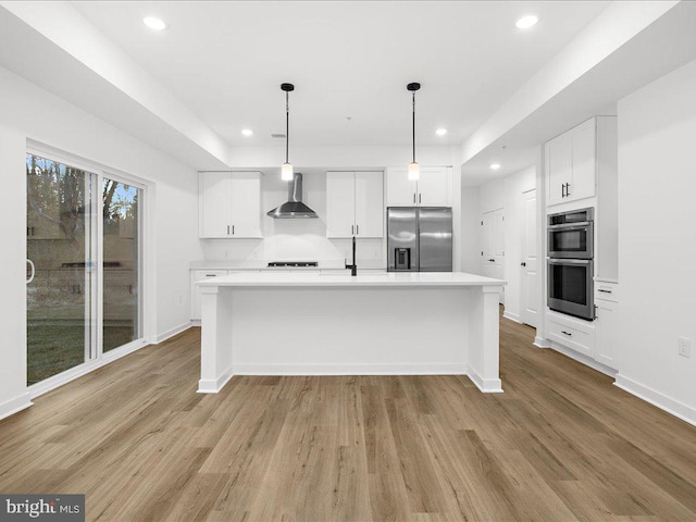 kitchen with appliances with stainless steel finishes, wall chimney range hood, pendant lighting, white cabinets, and an island with sink