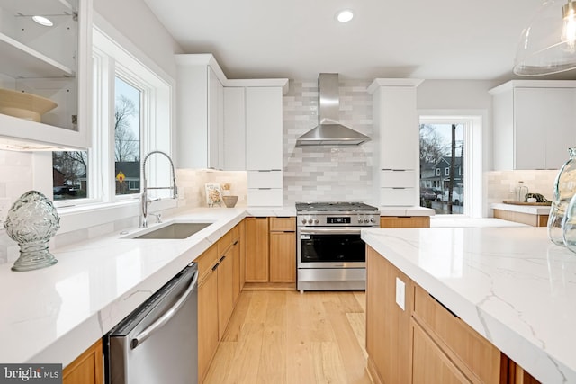 kitchen with light stone counters, stainless steel appliances, light wood-style floors, a sink, and wall chimney exhaust hood