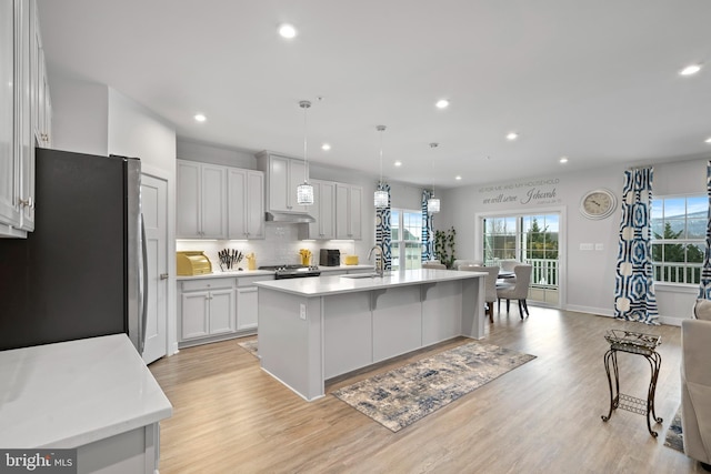 kitchen featuring stainless steel refrigerator, white cabinetry, light hardwood / wood-style flooring, hanging light fixtures, and an island with sink