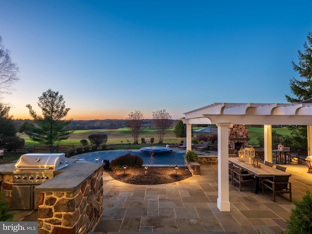 patio terrace at dusk featuring a grill, an outdoor stone fireplace, an outdoor kitchen, and a pool