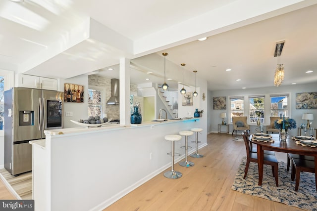 kitchen with stainless steel fridge with ice dispenser, decorative light fixtures, white cabinetry, kitchen peninsula, and wall chimney range hood