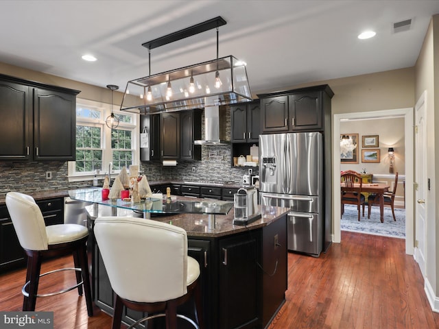 kitchen with stainless steel fridge, backsplash, wall chimney exhaust hood, a kitchen island, and hanging light fixtures