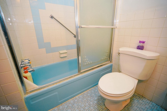 bathroom with tile patterned floors, toilet, bath / shower combo with glass door, and tile walls