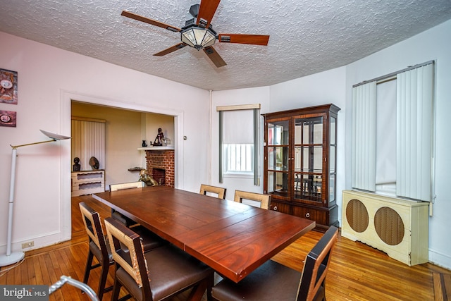 dining room featuring a brick fireplace, hardwood / wood-style flooring, a textured ceiling, and ceiling fan