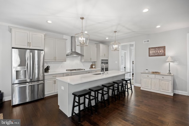 kitchen with wall chimney exhaust hood, hanging light fixtures, an island with sink, a kitchen bar, and appliances with stainless steel finishes