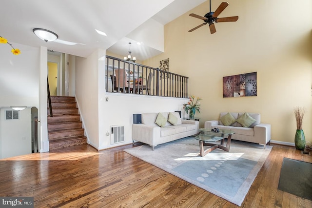 living room with ceiling fan with notable chandelier, a high ceiling, wood finished floors, visible vents, and stairs