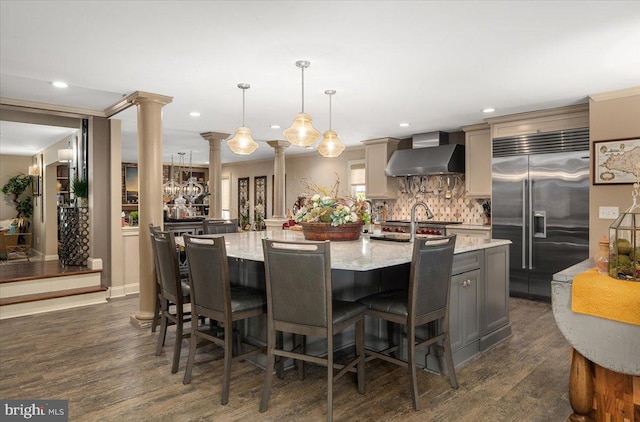 kitchen with light stone countertops, stainless steel built in refrigerator, wall chimney range hood, decorative light fixtures, and a center island with sink