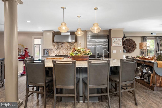 kitchen with a breakfast bar, stainless steel built in fridge, wall chimney range hood, hanging light fixtures, and an island with sink