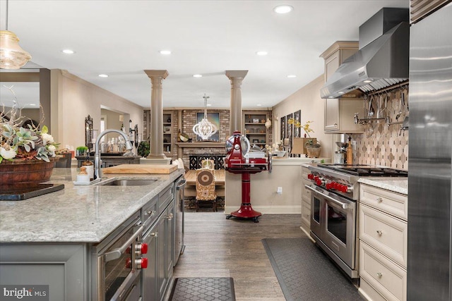 kitchen featuring decorative columns, light stone counters, stainless steel appliances, wall chimney range hood, and hanging light fixtures