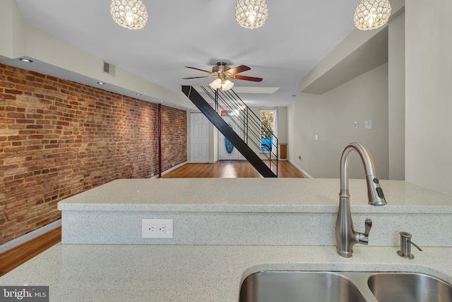 kitchen with light stone counters, sink, brick wall, and ceiling fan with notable chandelier