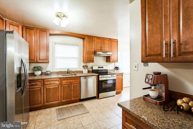 kitchen featuring brown cabinetry, stone counters, stainless steel appliances, under cabinet range hood, and a sink