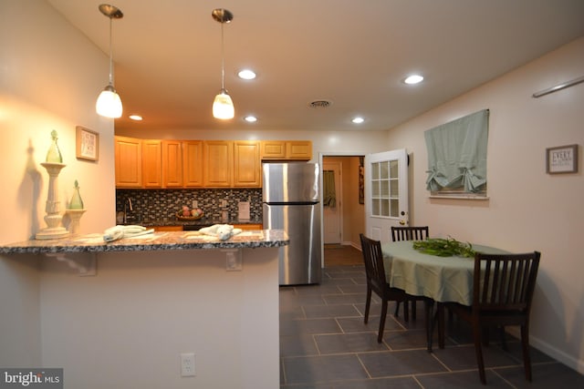 kitchen with hanging light fixtures, stainless steel fridge, light stone countertops, light brown cabinetry, and tasteful backsplash